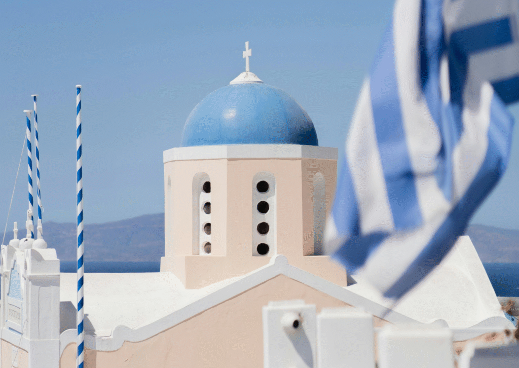 Charakterystyczna biała cerkiew z niebieską kopułą i grecką flagą – symbol architektury Cyklad, często spotykany na Santorini.