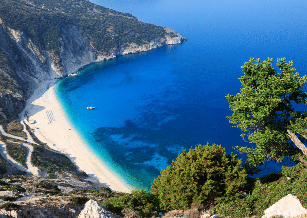 Widok z góry na plażę Myrtos na Kefalonii z jasnym piaskiem, turkusową wodą i żaglówką zacumowaną u brzegu.