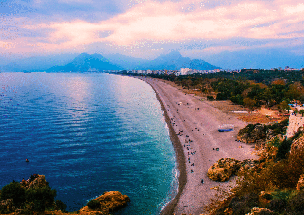 Panoramiczny widok na długą żwirową plażę Konyaaltı w Antalyi z górami i miastem w tle o zachodzie słońca.