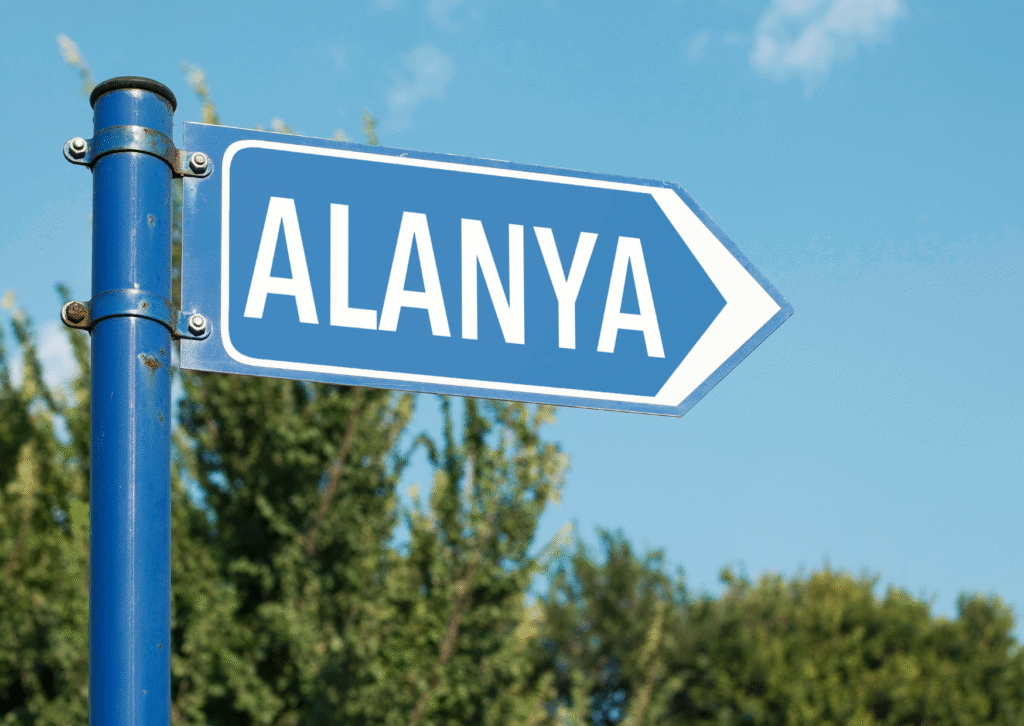 Niebieska tabliczka drogowa z napisem Alanya na tle błękitnego nieba i zielonych drzew.