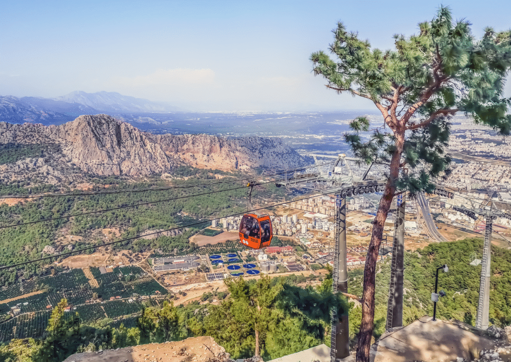 Czerwona gondola kolejki linowej Alanya Teleferik zawieszona nad miastem i górzystym krajobrazem.