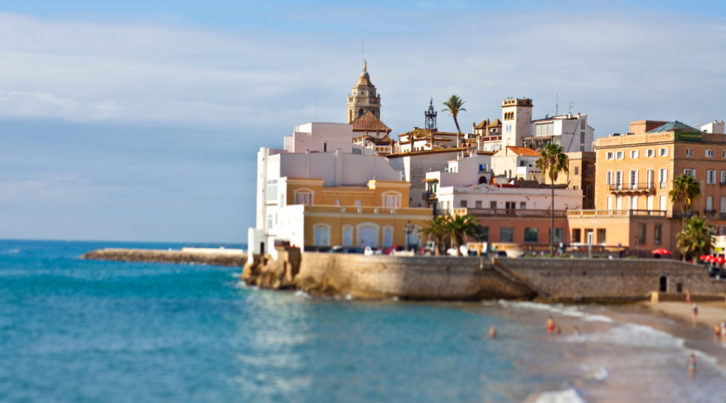 Kolorowe secesyjne budynki na skalistym cyplu nad lazurowym wybrzeżem Sitges, z palmami i wieżą kościoła w tle.