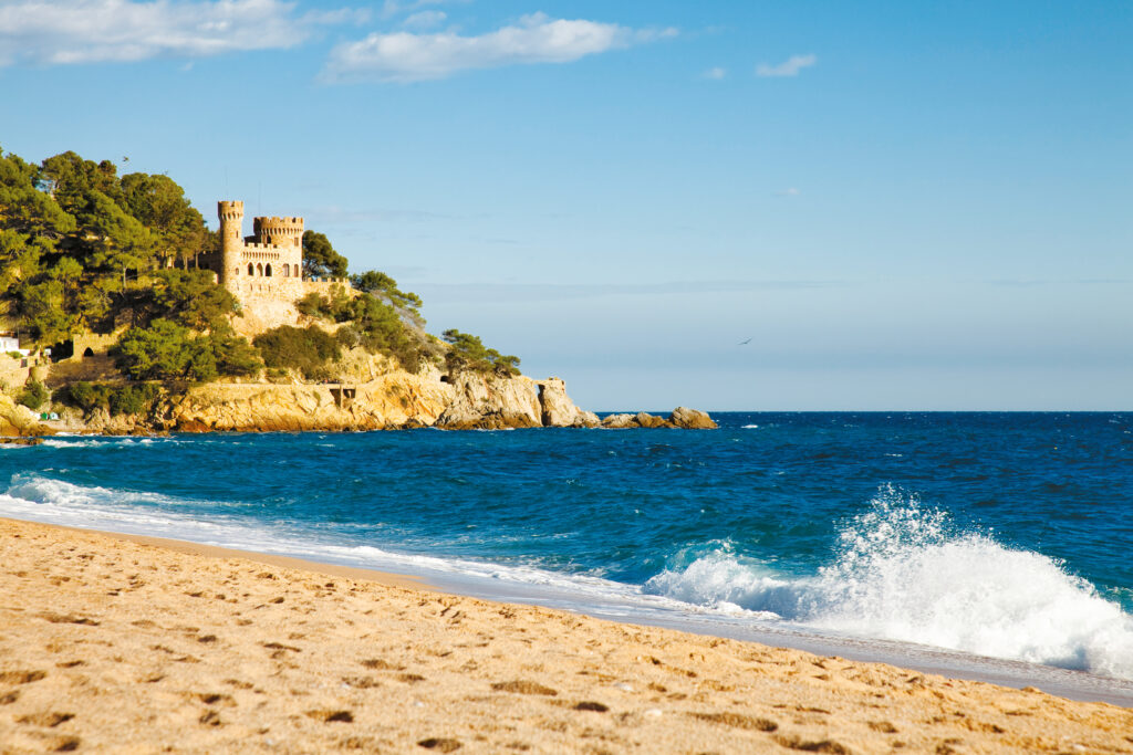 Widok na piaszczystą plażę Lloret de Mar z falującym Morzem Śródziemnym, a na skalistym zboczu malowniczy zamek Castell d’en Plaja.