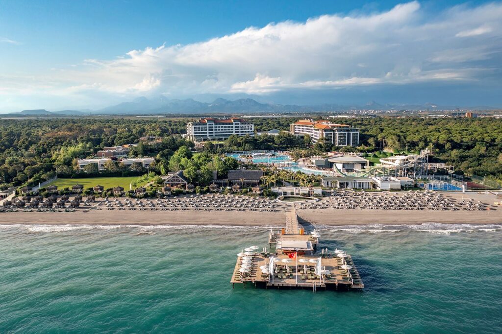 Widok z lotu ptaka na Ela Excellence Resort Belek – rozległy kompleks hotelowy otoczony lasem piniowym, z basenami, zjeżdżalniami wodnymi, prywatną plażą z pomostem i licznymi leżakami.