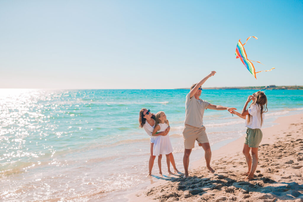 Rodzina na plaży – ojciec puszcza latawiec, mama obejmuje córkę, a starsza córka patrzy na latawiec nad turkusową wodą.