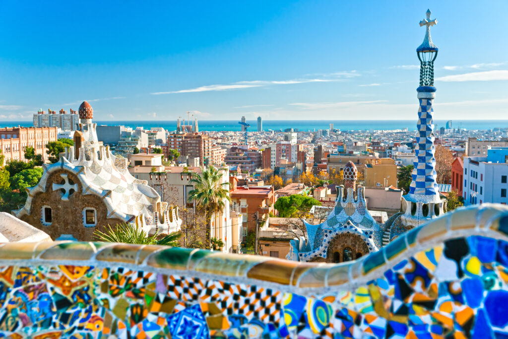 Panoramiczny widok z Parku Güell w Barcelonie: w pierwszym planie kolorowa mozaikowa ławka Gaudíego, w tle rozległa zabudowa miasta z widoczną katedrą Sagrada Família nad horyzontem Morza Śródziemnego.