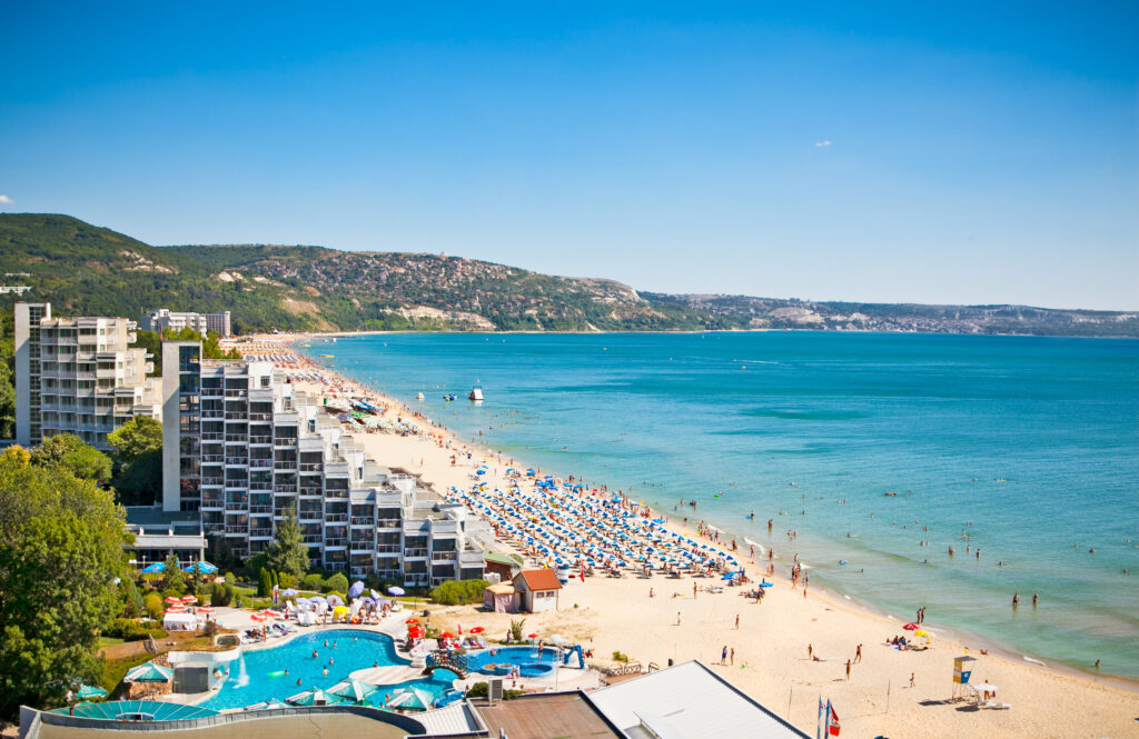 Widok na piaszczystą plażę nad Morzem Czarnym w Bułgarii, z leżakami, parasolami i turkusową wodą, w tle hotele na wybrzeżu.
