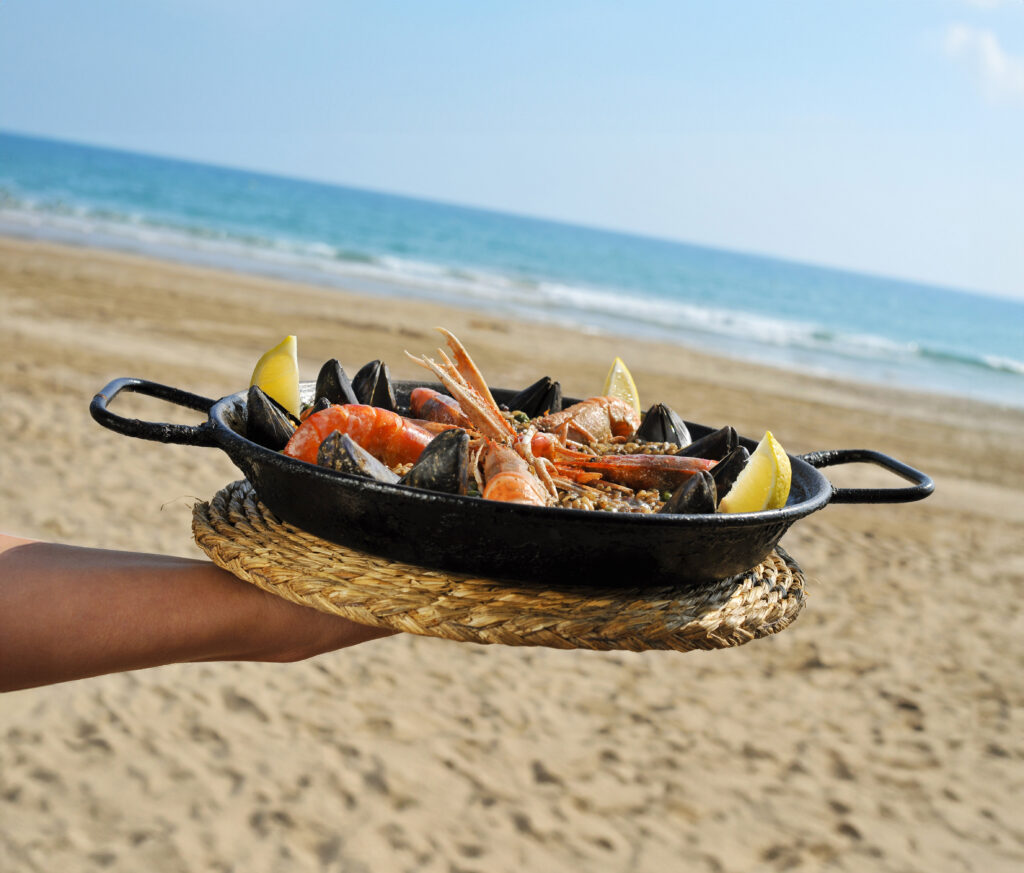 Patelnia z paellą z owocami morza (krewetki, mule), podana na plaży z widokiem na morze.