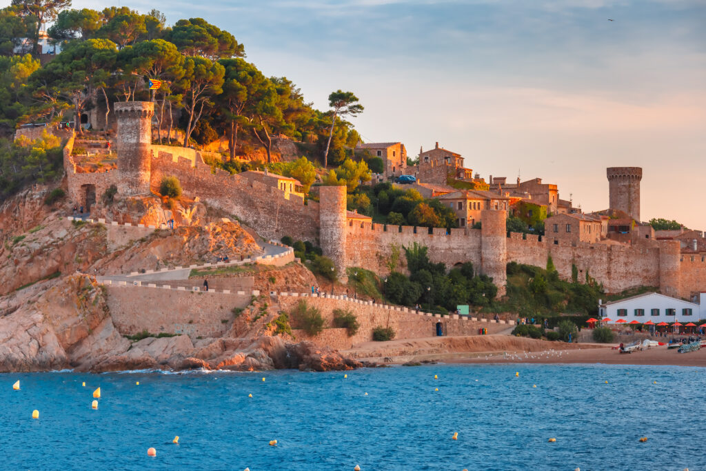 Zachód słońca nad nadmorskim miasteczkiem Tossa de Mar na Costa Brava w Hiszpanii – warowny mur zamku, piaszczysta plaża i spokojne wody Morza Śródziemnego.