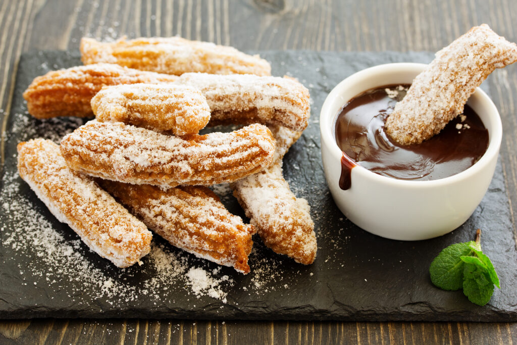 Kilka podłużnych churros oprószonych cukrem i posypką, obok miseczka gęstej, czekoladowej polewy.