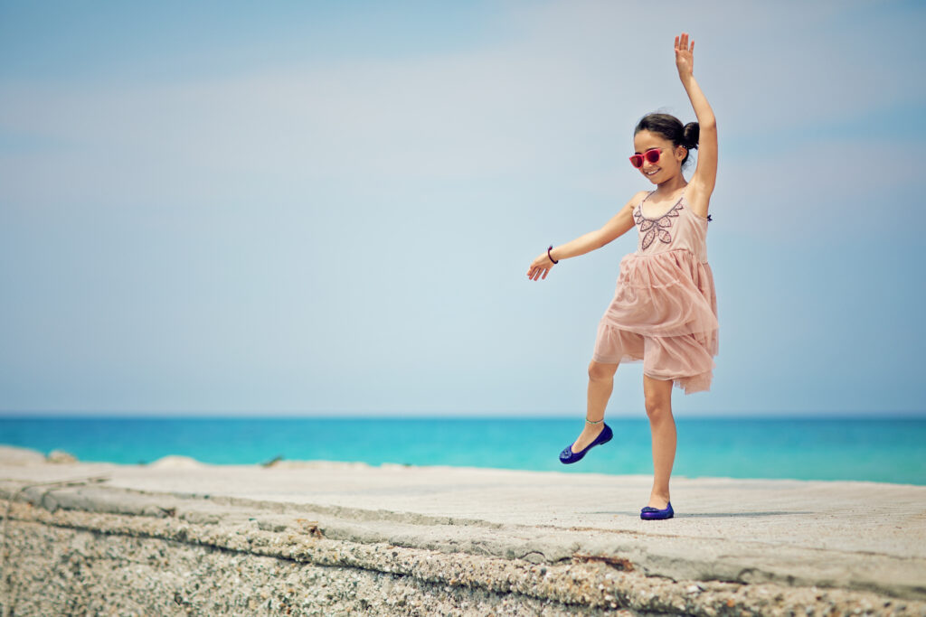 Uśmiechnięta dziewczynka w różowej sukience i czerwonych okularach przeciwsłonecznych, bawiąca się na kamiennym nabrzeżu z turkusową wodą w tle.