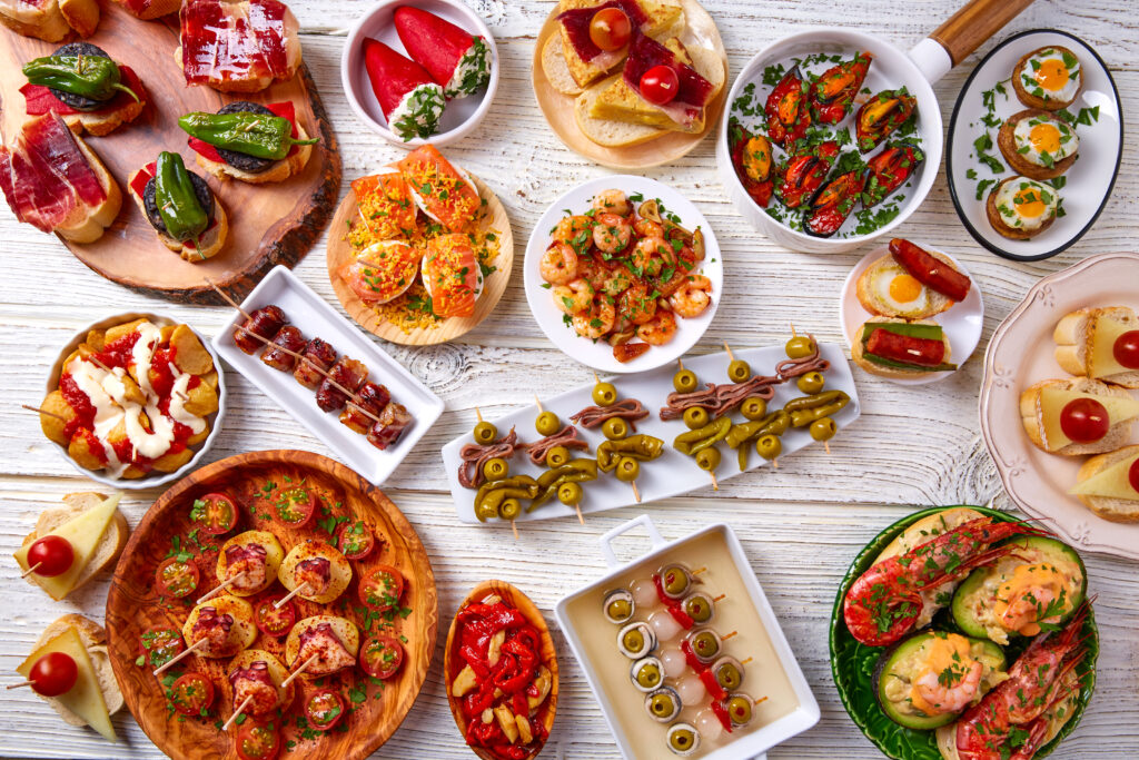 Stół zastawiony różnorodnymi hiszpańskimi tapas: oliwki na wykałaczkach, krewetki, pieczone papryki, mini kanapki z szynką, patatas bravas i inne przekąski