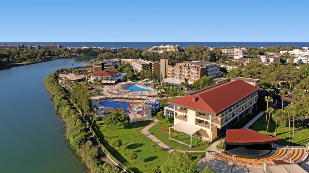 Widok z drona na Marvida Family Eco Resort – kompleks niskich budynków wokół dwóch basenów, z aquaparkiem dla dzieci, położony nad spokojnym jeziorem i wśród zieleni, z morzem w tle.