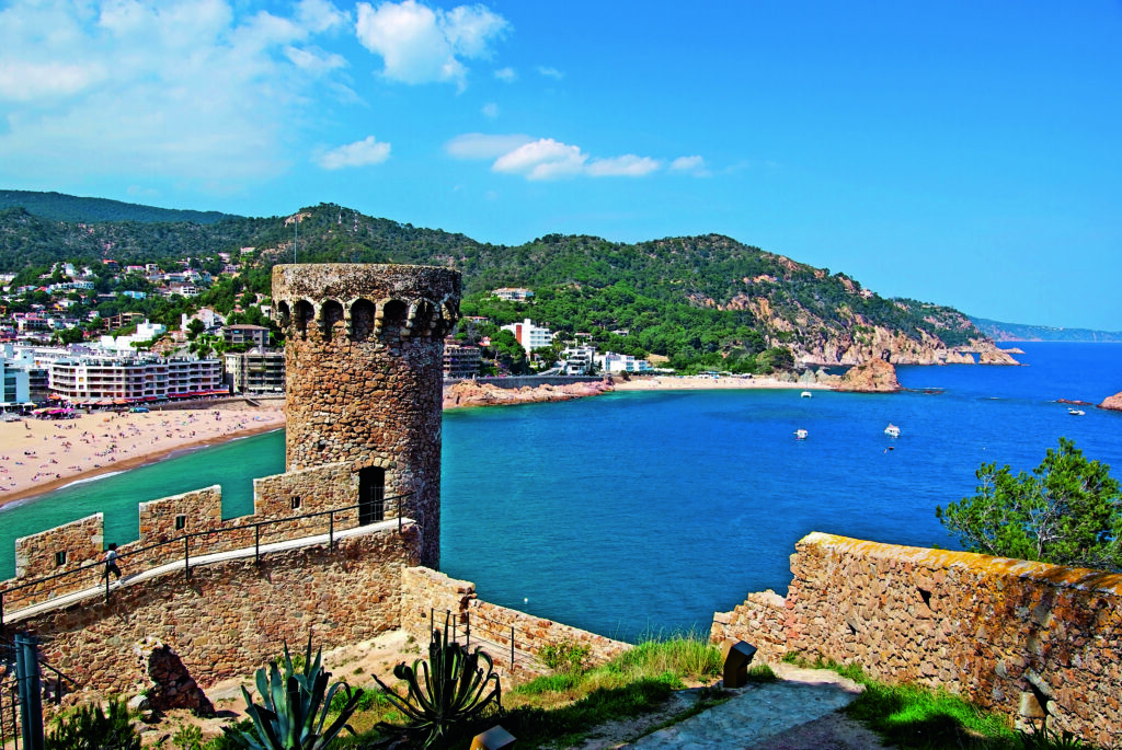 Panoramiczny widok na średniowieczne mury i wieżę zamku w Tossa de Mar, z piaszczystą plażą i lazurowym Morzem Śródziemnym w tle.