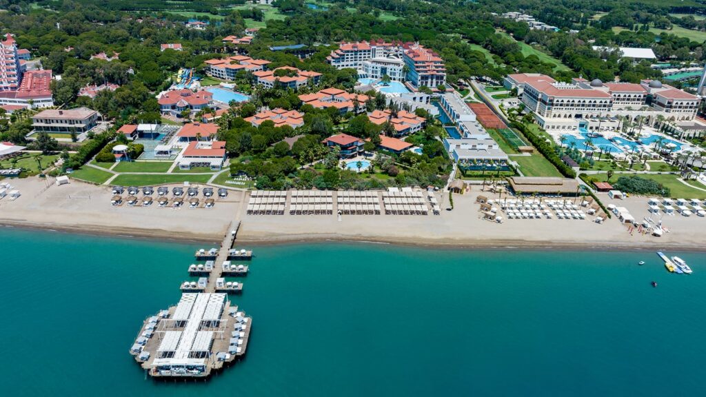 Panoramiczny widok z drona na rozległy kompleks Sirene Belek Hotel nad brzegiem morza, z prywatnym pomostem, plażą ze słonecznikami oraz basenami otoczonymi hotelowymi budynkami i bujną zielenią.