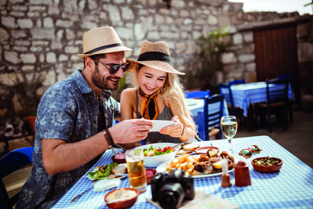 Uśmiechnięta para turystów podczas urlopu w Grecji, jedząca lunch w tradycyjnej greckiej restauracji i przeglądająca zdjęcia na smartfonie.