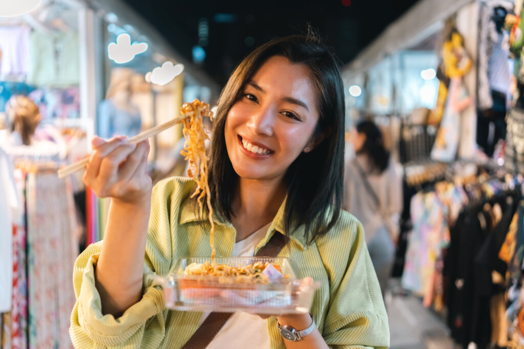 Kobieta jedząca pad thai na ulicznym targu nocnym w Tajlandii, trzymająca makaron pałeczkami i uśmiechająca się do kamery