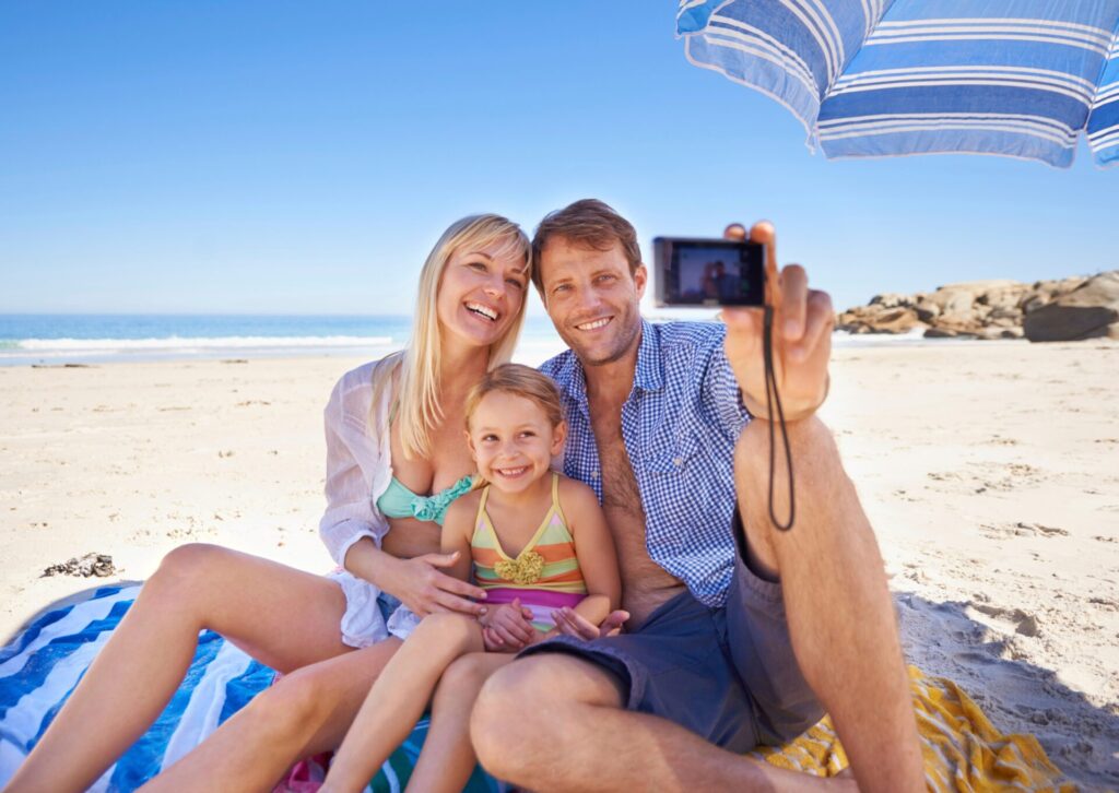 Uśmiechnięte małżeństwo z małą córką siedzące na piaszczystej plaży pod niebiesko-białym parasolem i robiące wspólne zdjęcie aparatem cyfrowym.