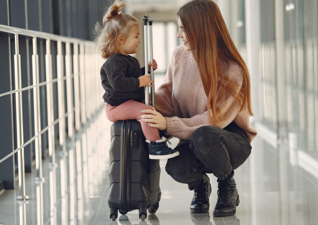 Uśmiechnięta mama kucająca obok małej córeczki siedzącej na czarnej walizce kabinowej na lotnisku