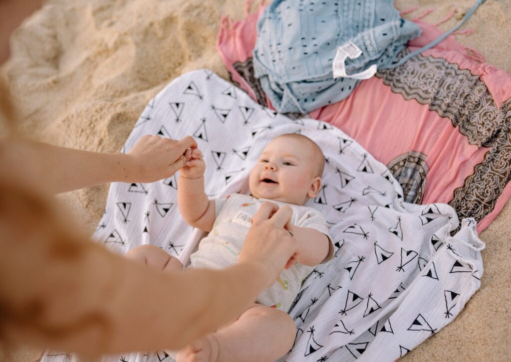 Uśmiechnięte niemowlę leżące na białym kocyku w czarne wzory na piaszczystej plaży i trzymające rodzica za dłonie.