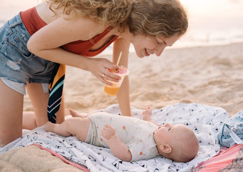 Uśmiechnięta kobieta w jeansowych szortach trzymająca napój z rurką i patrząca na niemowlę leżące na kocu na plaży.
