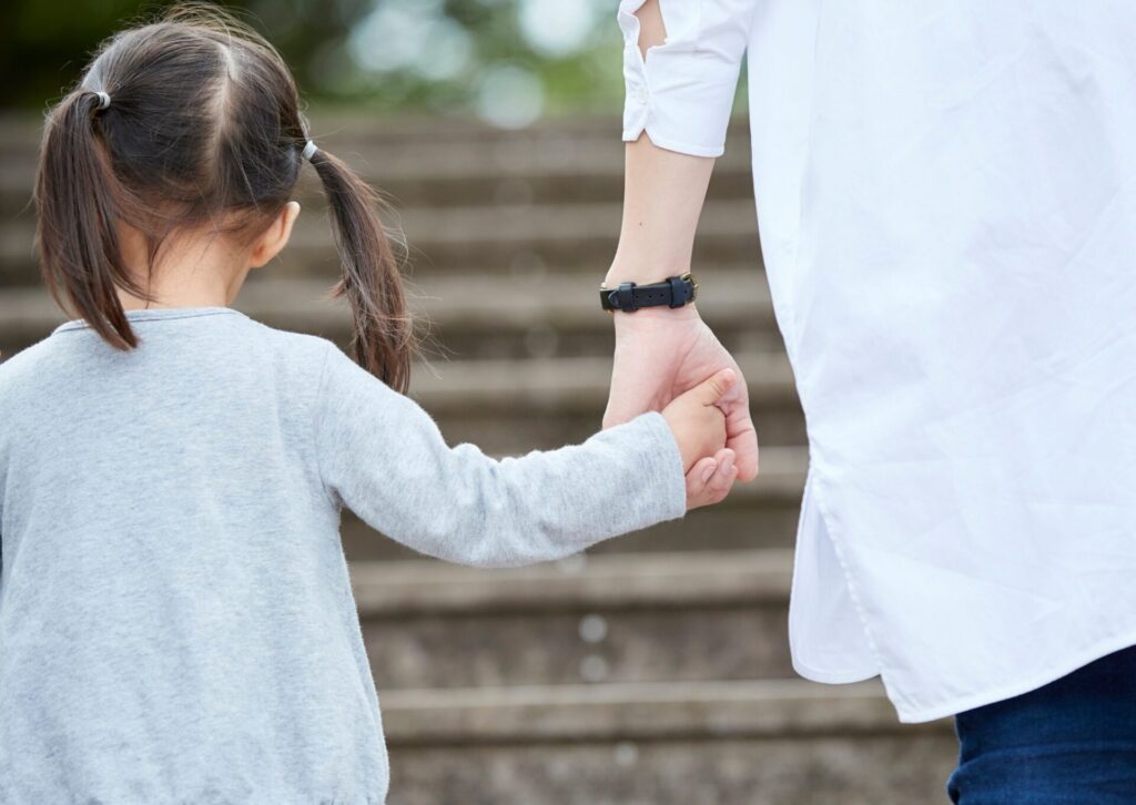 Mała dziewczynka w szarej bluzce z dwoma kucykami idąca po schodach i trzymająca za rękę rodzica w białej koszuli.