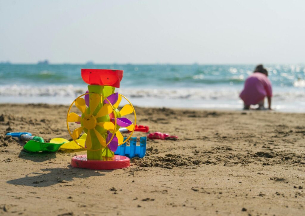 Kolorowy młynek wodny do piasku, łopatki i grabki leżące na plaży przed kucającym nad brzegiem morza dzieckiem