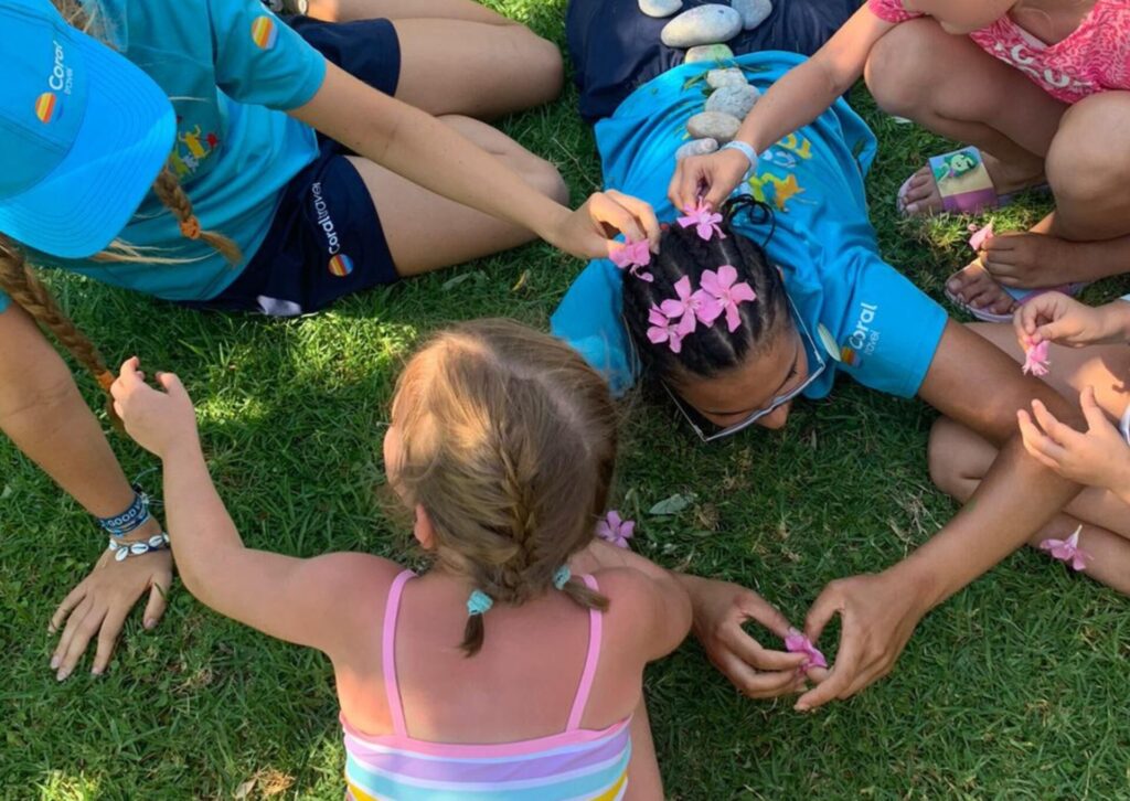 Dzieci i animatorki Coral Family Club w koszulkach Coral Travel bawią się na trawie, wpinając różowe kwiaty we włosy podczas animacji