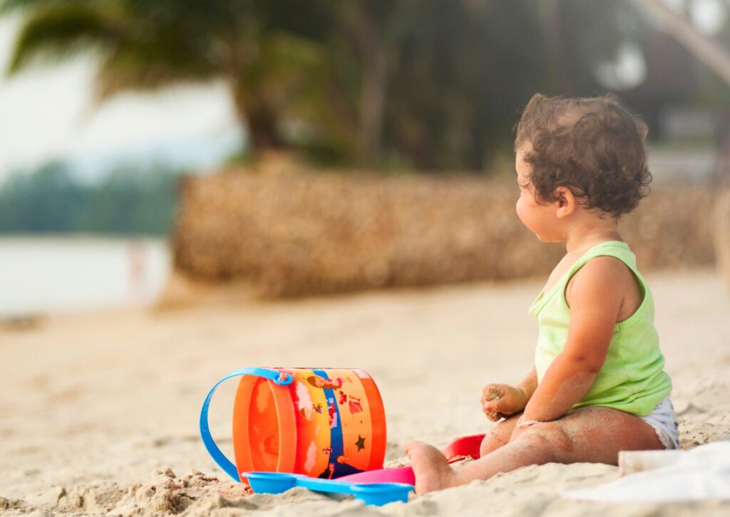 Małe dziecko w zielonej koszulce siedzące tyłem na piaszczystej plaży obok kolorowego wiaderka i łopatki do piasku