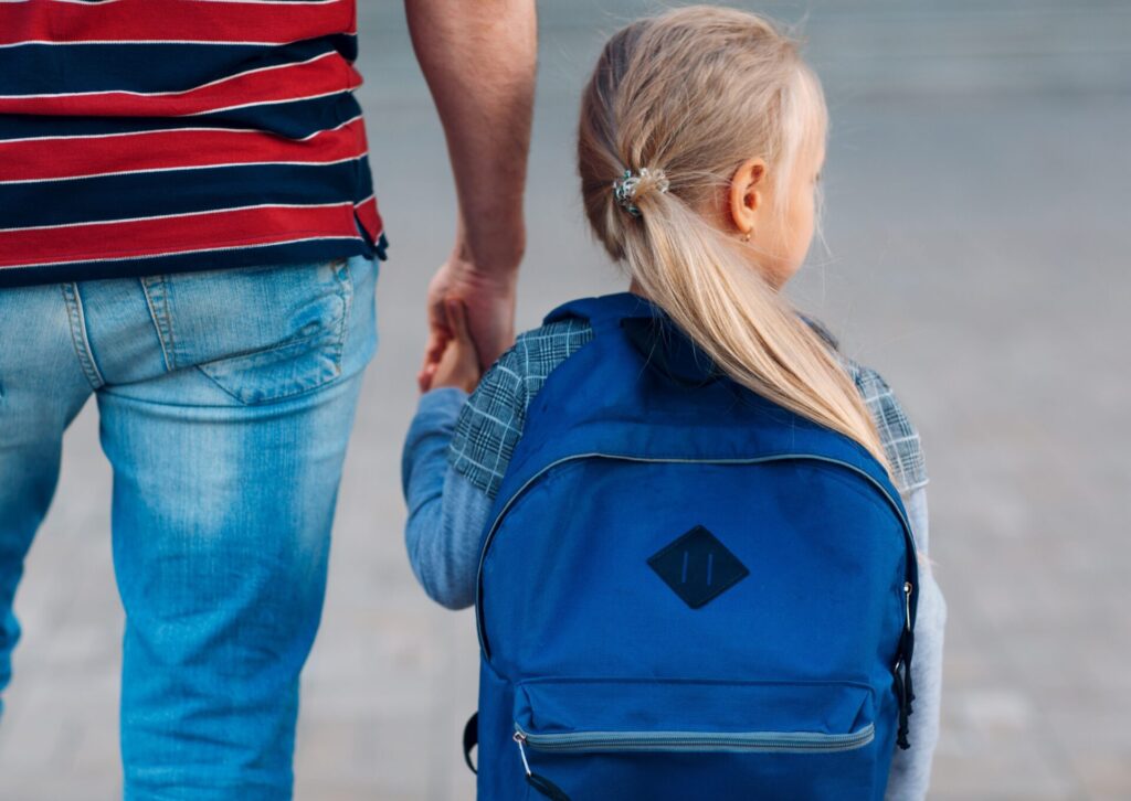 Dziewczynka z długimi blond włosami w kucyku i z dużym niebieskim plecakiem trzymająca za rękę rodzica w jeansach i pasiastej koszulce.