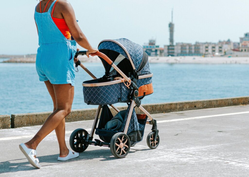 Kobieta w jeansowych ogrodniczkach prowadząca pikowany wózek dziecięcy po nadmorskiej promenadzie podczas wakacji.