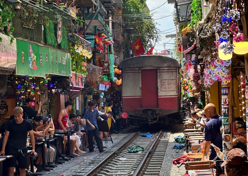 Gwarny i wąski przesmyk Train Street w Hanoi z kawiarniami po obu stronach torów, gdzie siedzą liczni turyści, a w głębi widać przejeżdżający tył pociągu