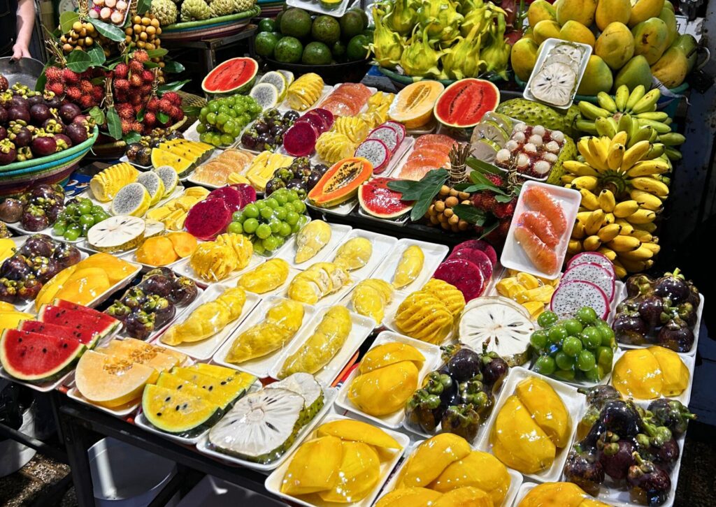 Bogato zaopatrzone stoisko targowe pełne świeżych, egzotycznych owoców, takich jak pokrojone mango, arbuzy, smocze owoce, banany, marakuja, mangostan, durian i rambutan, ułożonych na tackach i w koszach