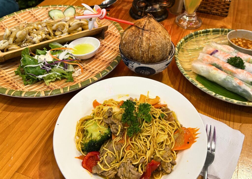 Tradycyjne wietnamskie potrawy podane na drewnianym stole, w tym smażony makaron z wołowiną i brokułami na talerzu, sajgonki bez smażenia (spring rolls), grillowane kalmary na bambusowej tacy oraz świeży kokos do picia ze słomką